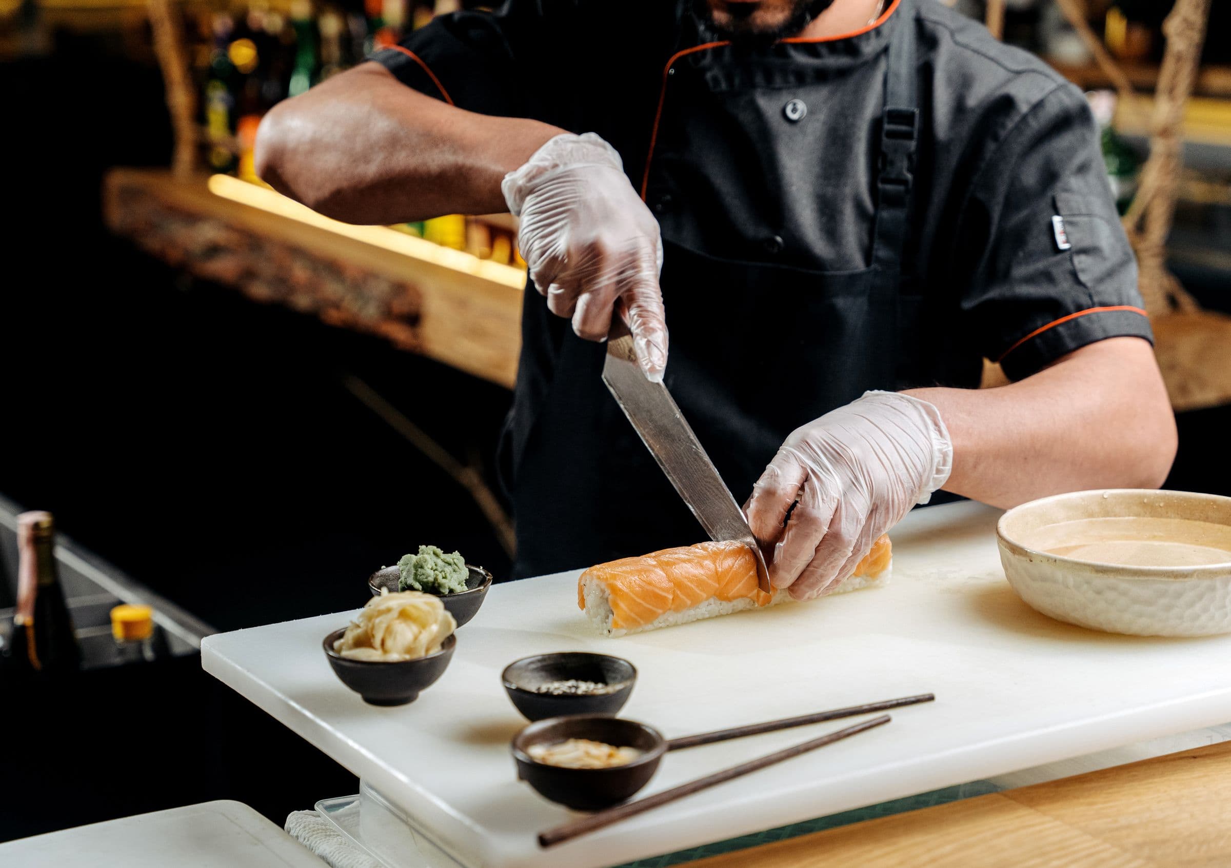 A chef slicing a salmon sushi roll with a long Japanese slicer on a white board